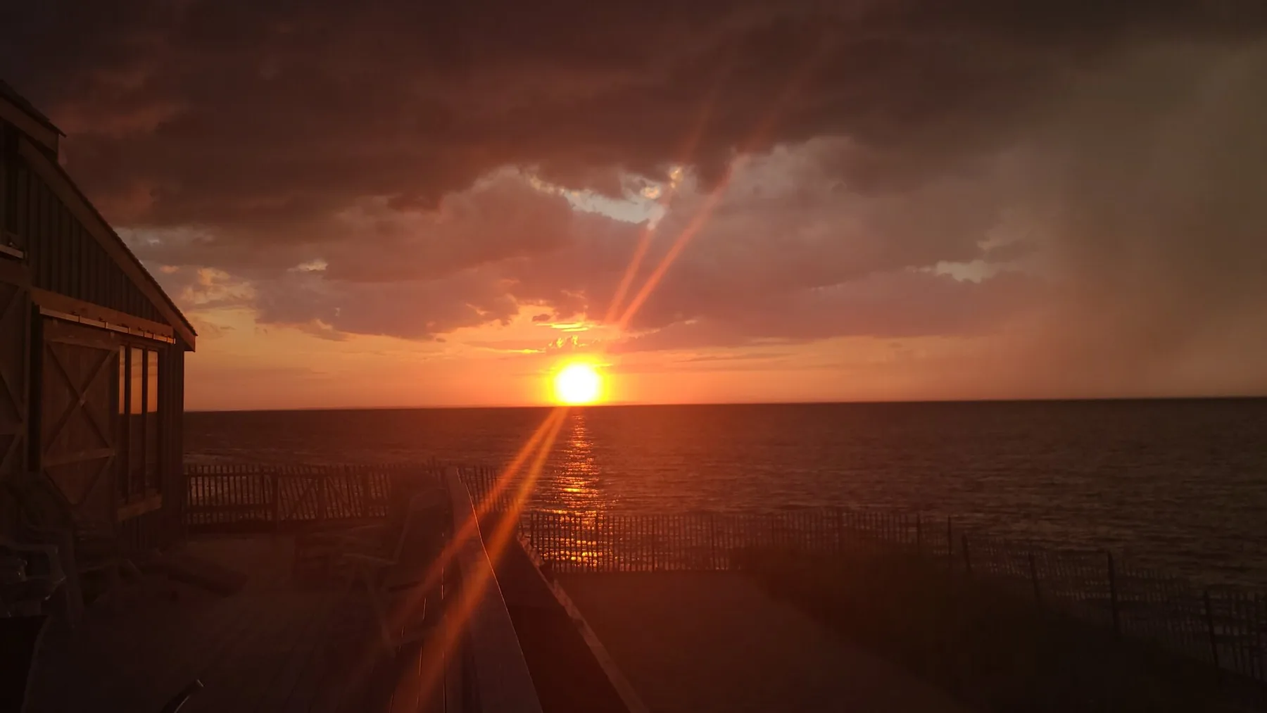 The sun sets over the ocean beside a wooden deck.