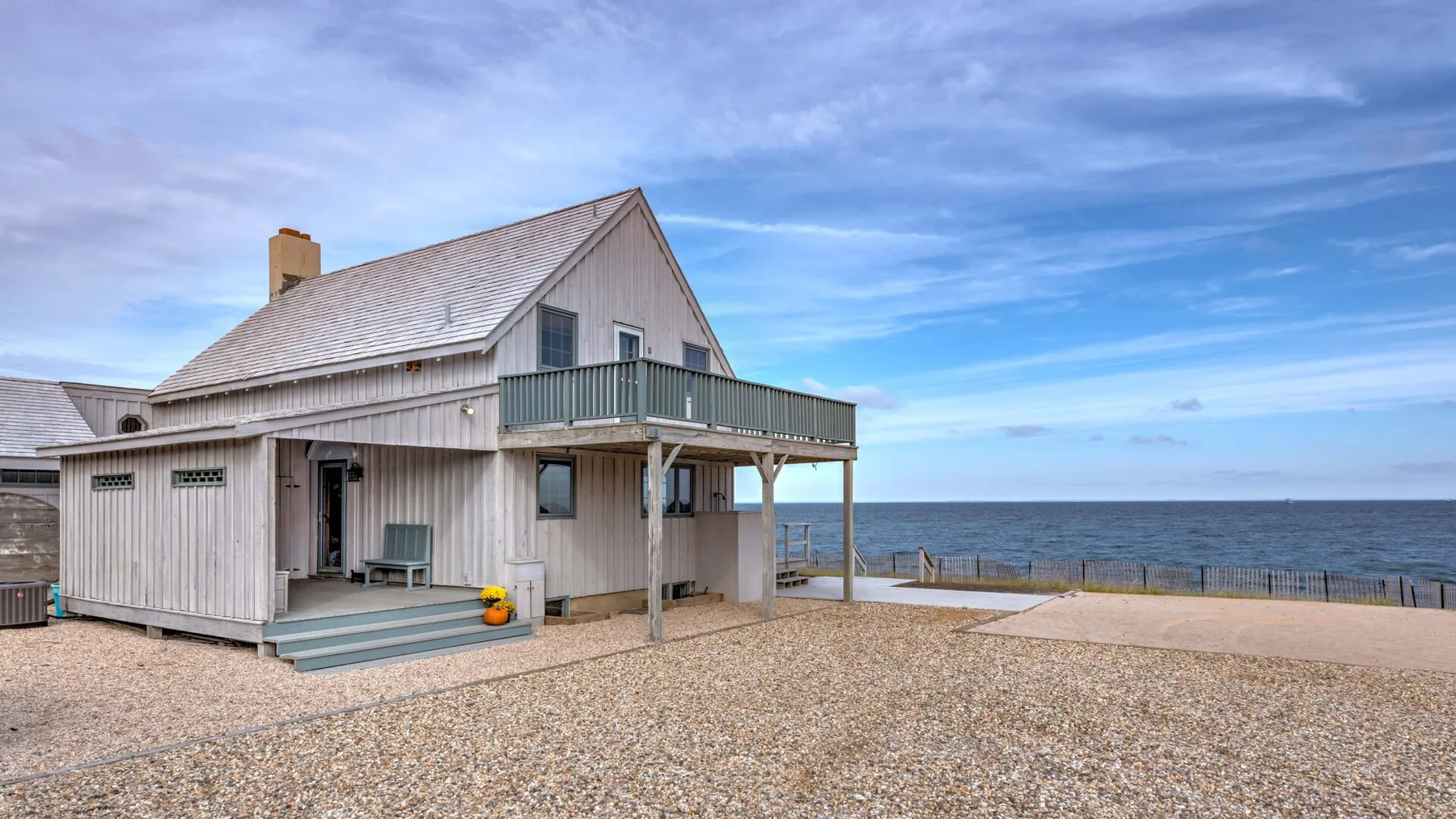 A raised seaside house with a deck overlooks a pebbled shore.