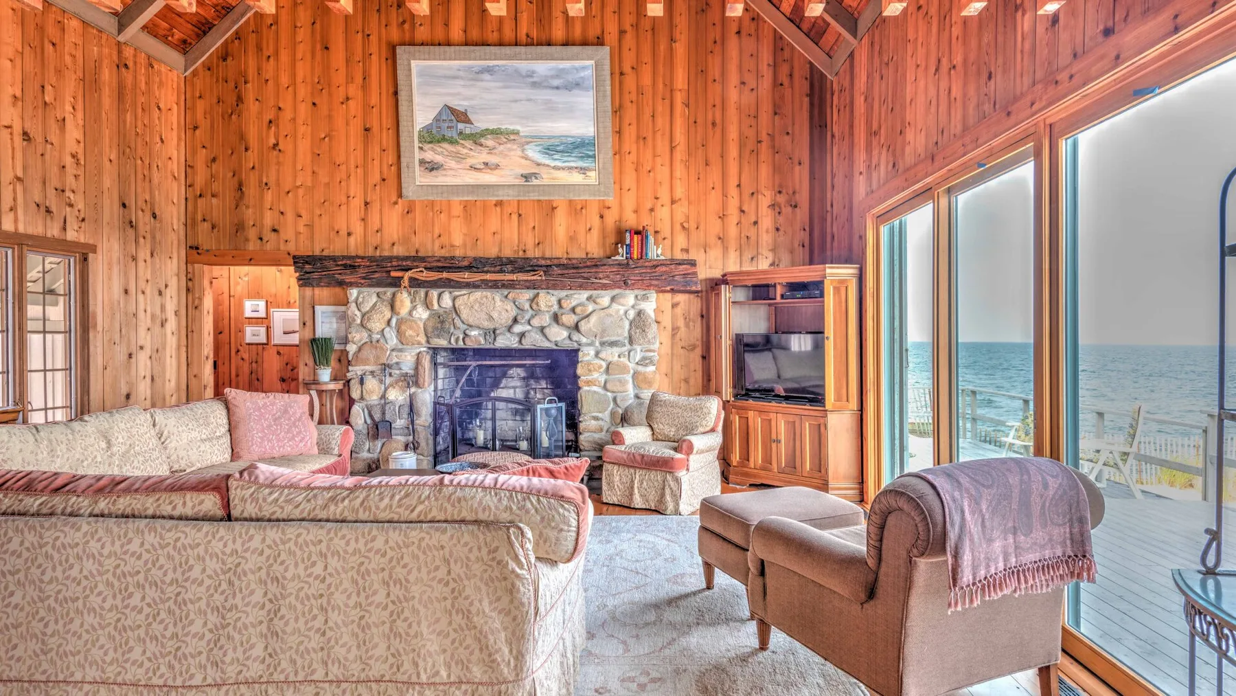 A wood-paneled living room with a stone fireplace and large windows overlooking the water.