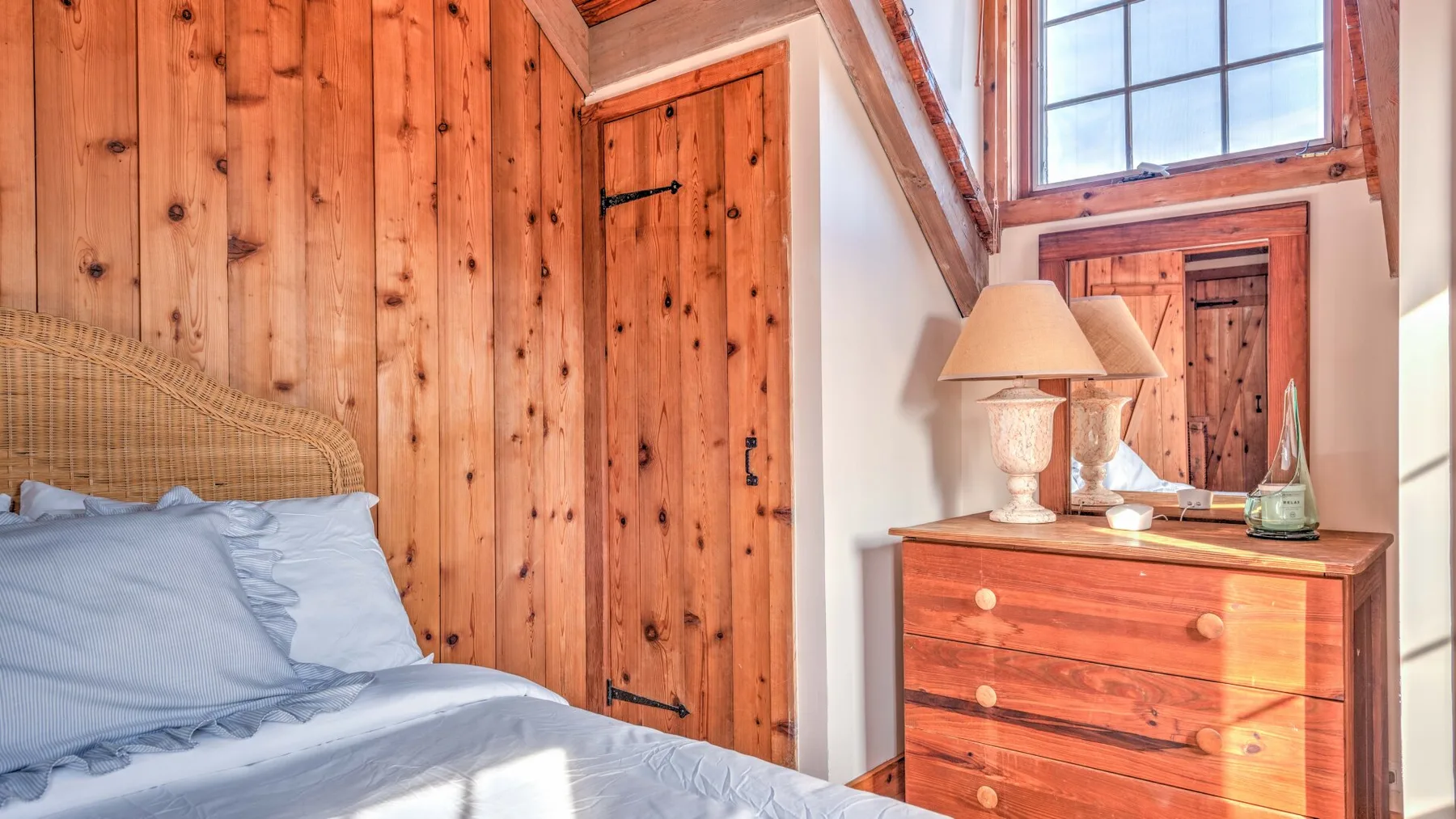 A bedroom with a bed, wooden walls, and a dresser beneath a bright window.