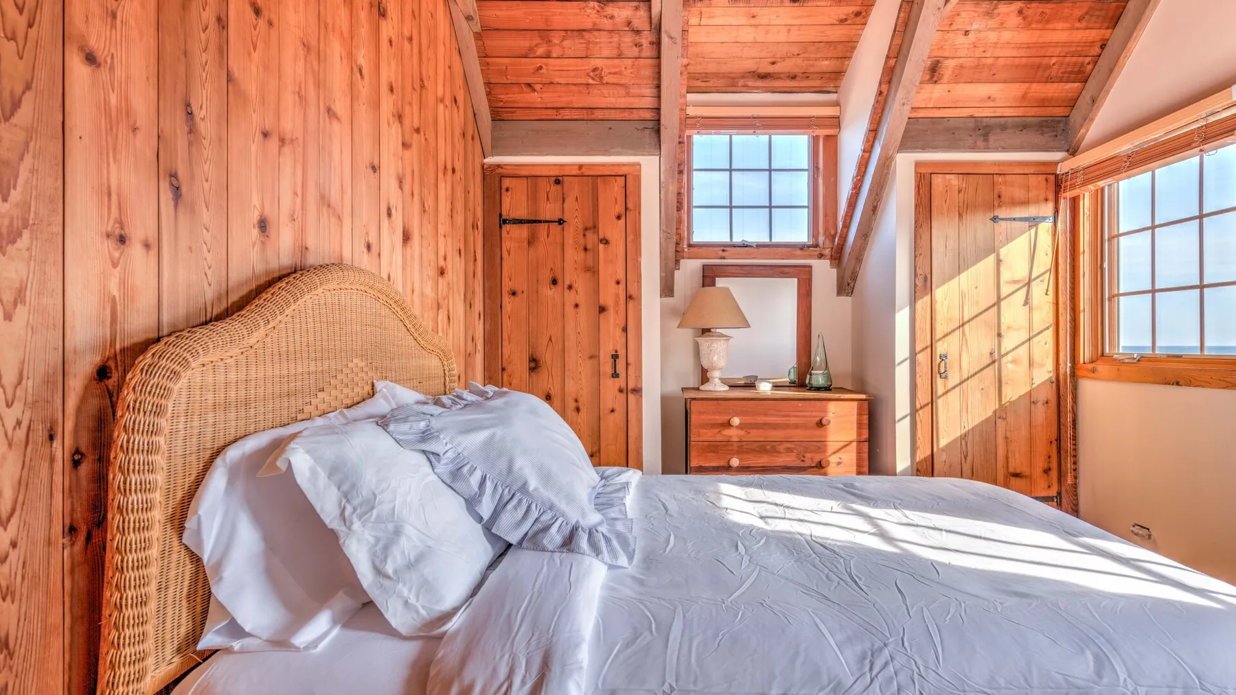 A bedroom with a bed beneath a sloped wooden ceiling and bright windows.