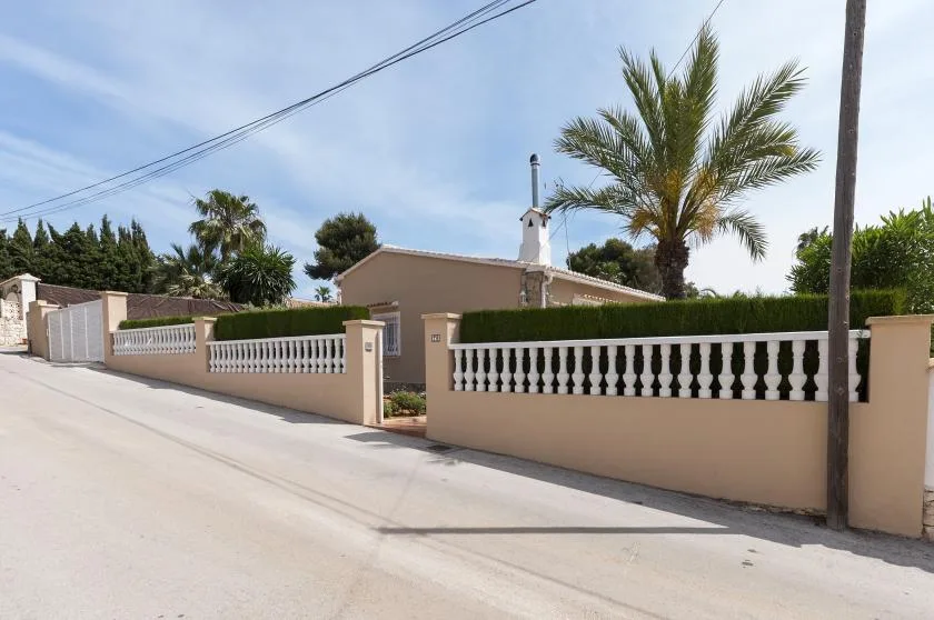 Una calle residencial con un muro bajo, una entrada con portón y palmeras.