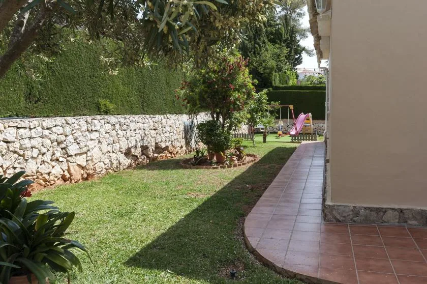 Un césped ajardinado junto a un muro de piedra, con un sendero pavimentado junto a la casa.