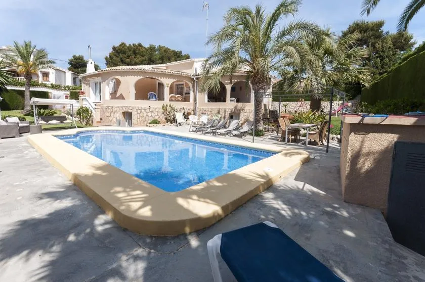 Una piscina está en un patio soleado con palmeras y una terraza cubierta.