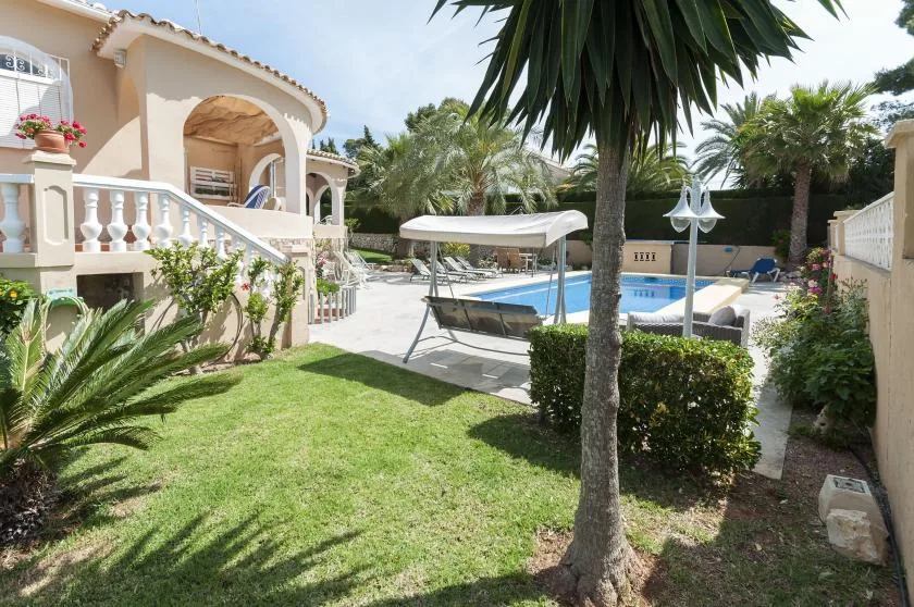 Un jardín rodea una piscina junto a una villa clara con balcones arqueados.