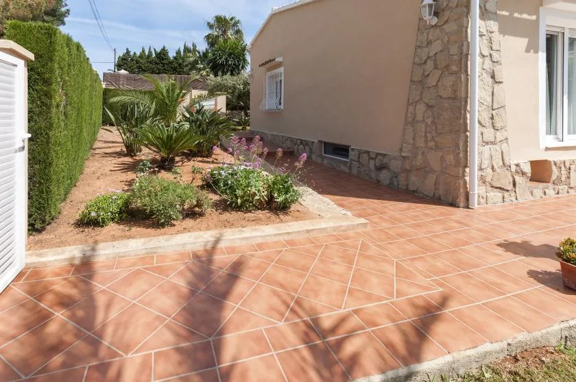 Un patio de baldosas junto a una casa, con un jardín y setos altos.
