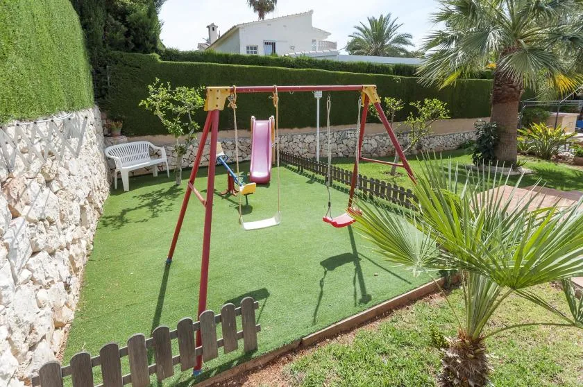 Zona de juegos infantil con un tobogán y columpios sobre césped artificial.