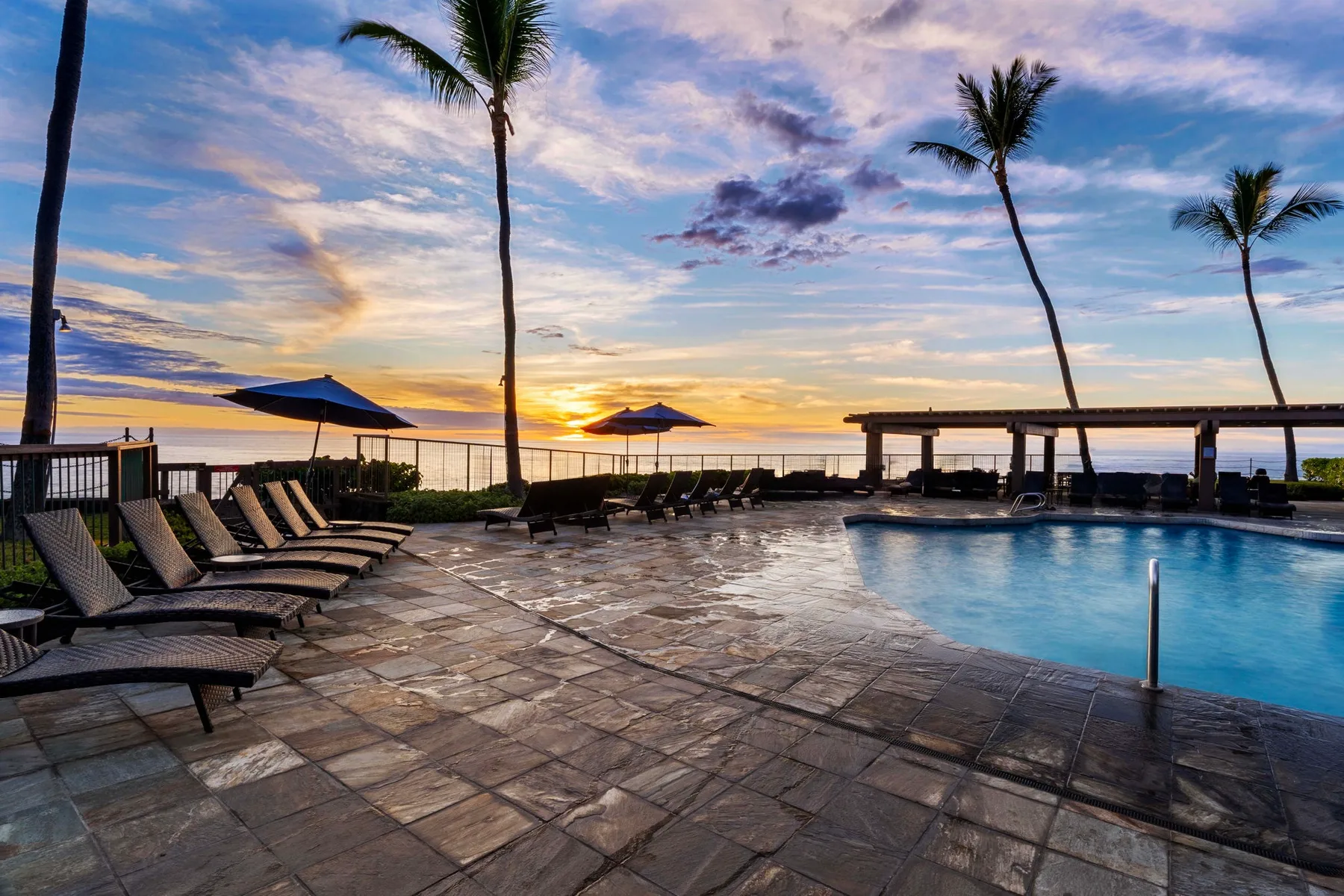 Un patio junto a la piscina mira al océano al atardecer, con asientos y palmeras.
