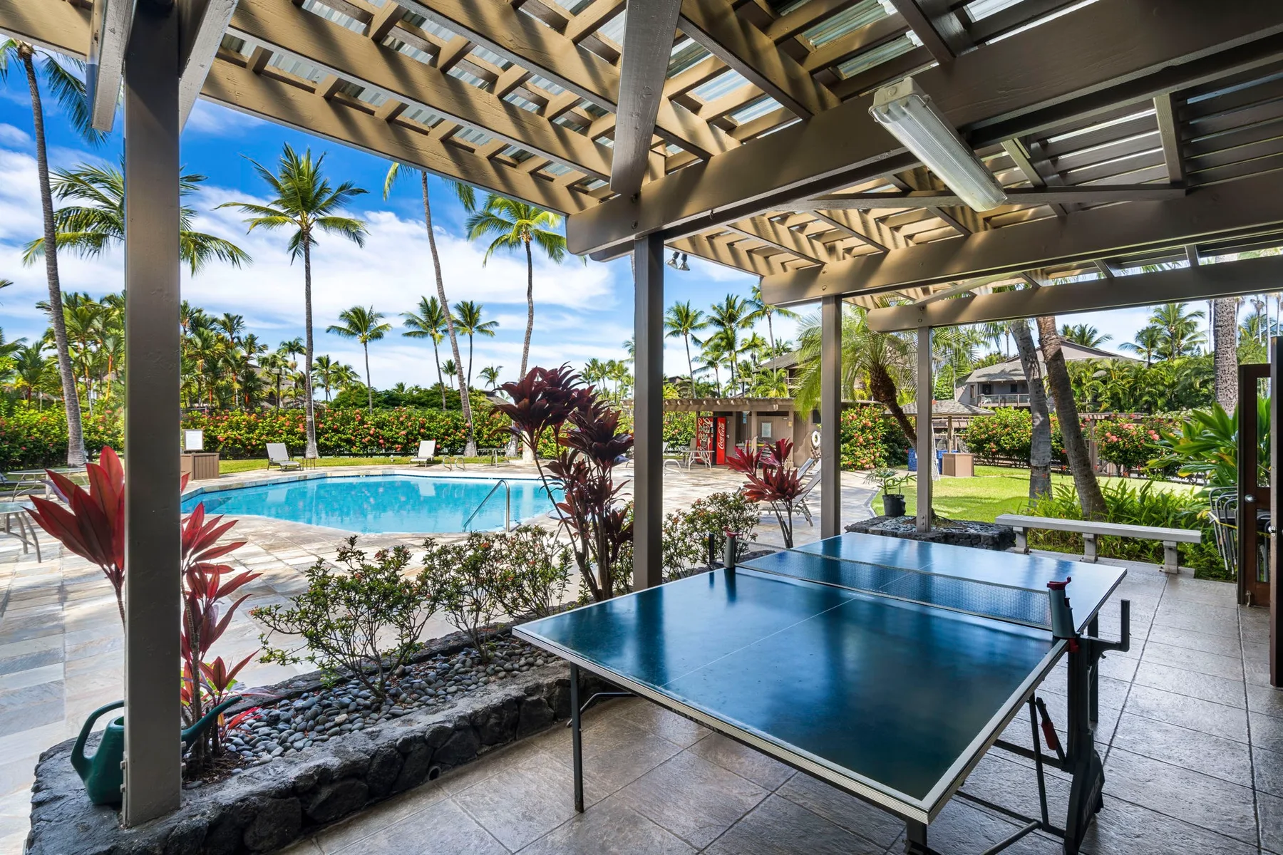 Una mesa de ping-pong está bajo un patio cubierto, junto a una piscina y vegetación tropical.