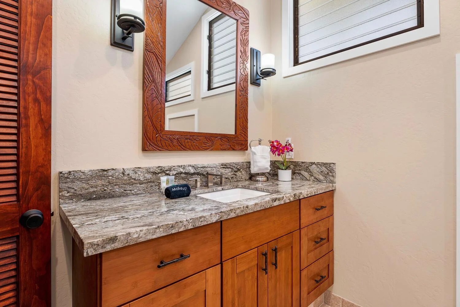 Un tocador de baño con encimera de piedra, espejo y un pequeño arreglo floral.