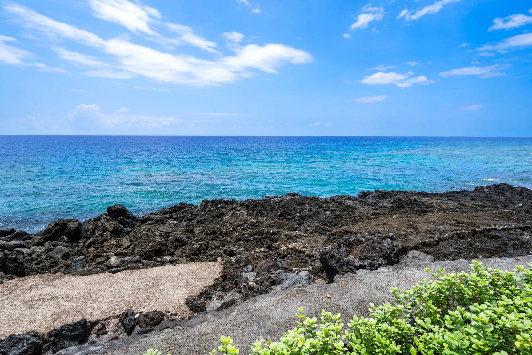 A rocky shoreline borders clear blue ocean water beneath a partly cloudy sky.