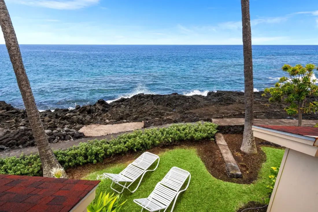 A grassy seaside patio with lounge chairs overlooks rocky shore and ocean waves.