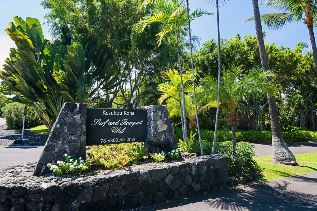A landscaped entrance features a stone sign surrounded by tropical plants.