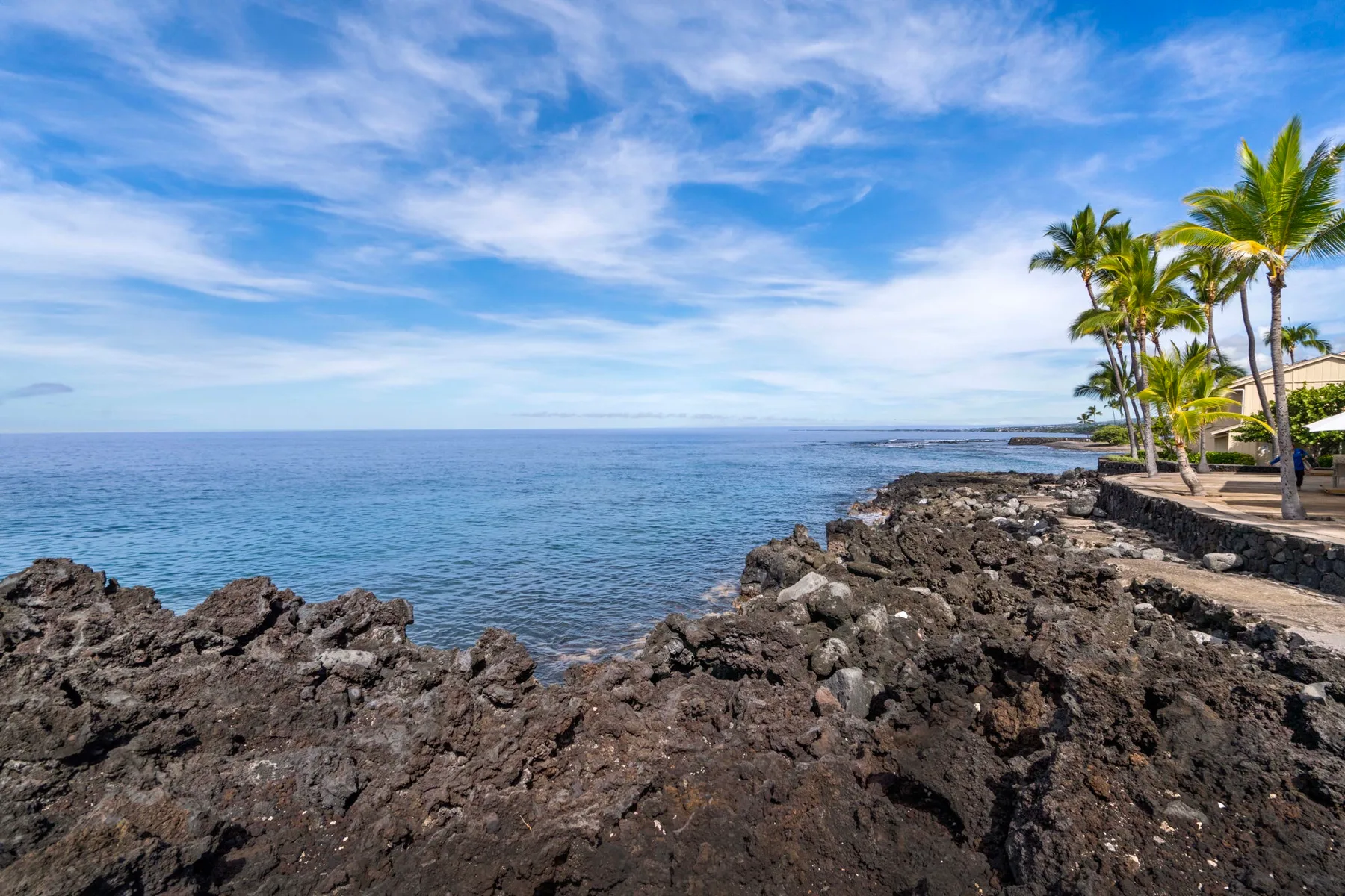 A rocky shoreline borders calm blue ocean water beneath a partly cloudy sky.