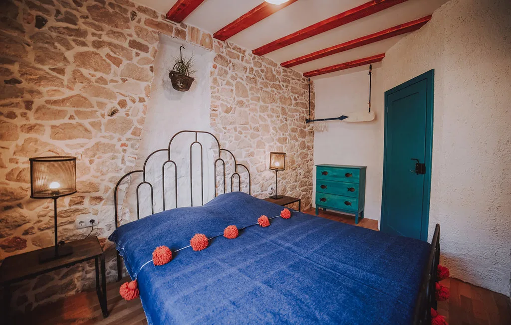 Un dormitorio con una colcha azul, paredes de piedra y una puerta verde azulado.