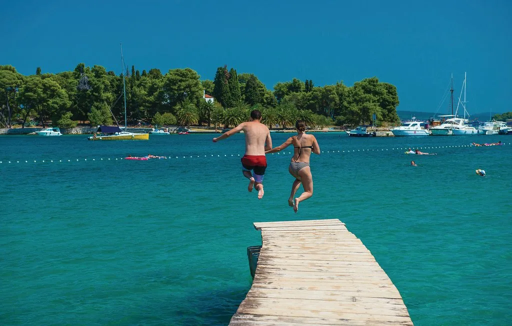 Dos personas saltan desde un muelle de madera a aguas azules.