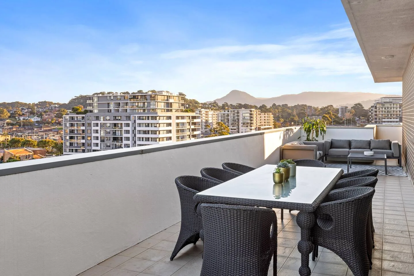 An outdoor balcony dining area has a table, seating, and views of nearby buildings and hills.