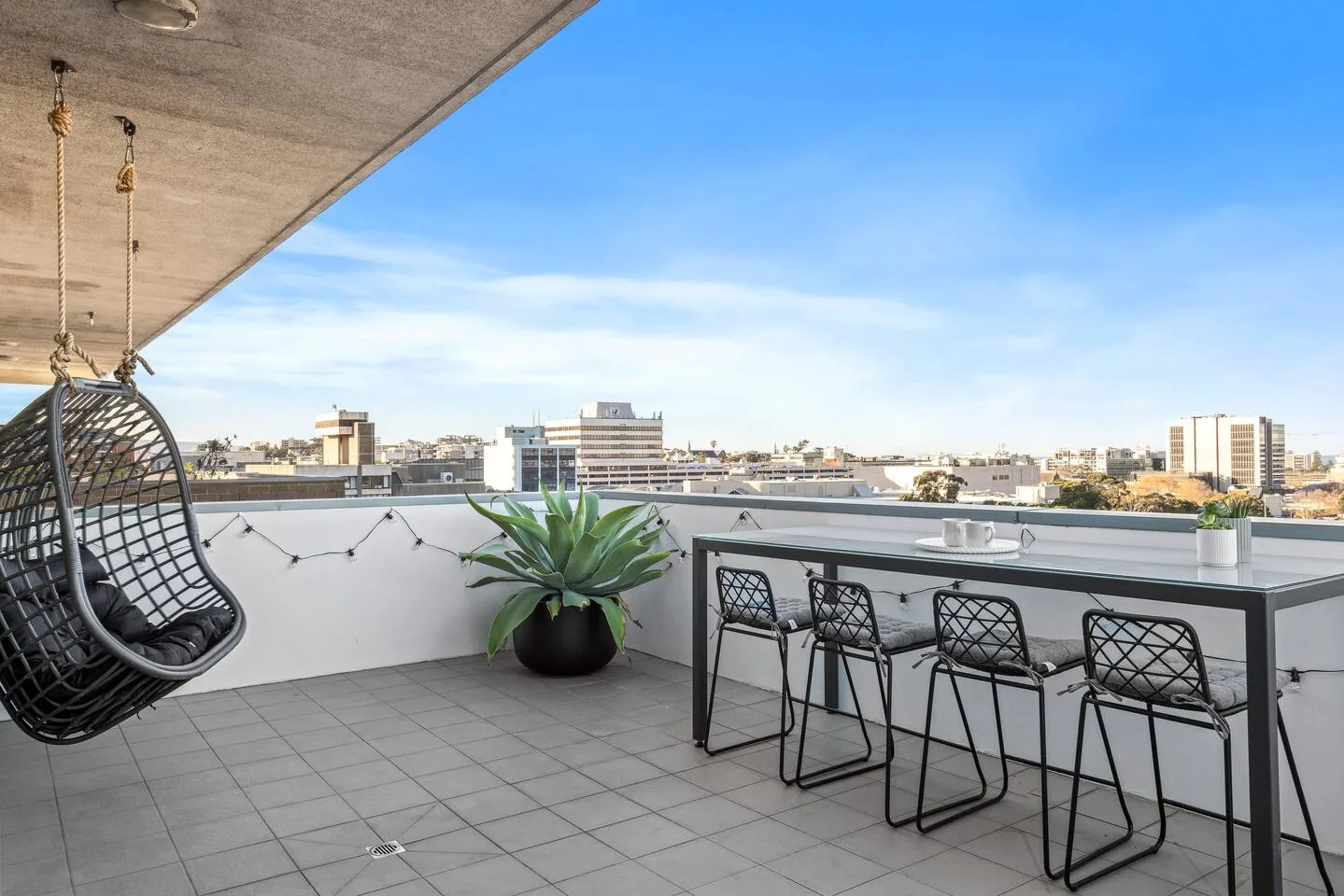 A rooftop terrace has a hanging seat, potted plant, and bar-height table with stools.