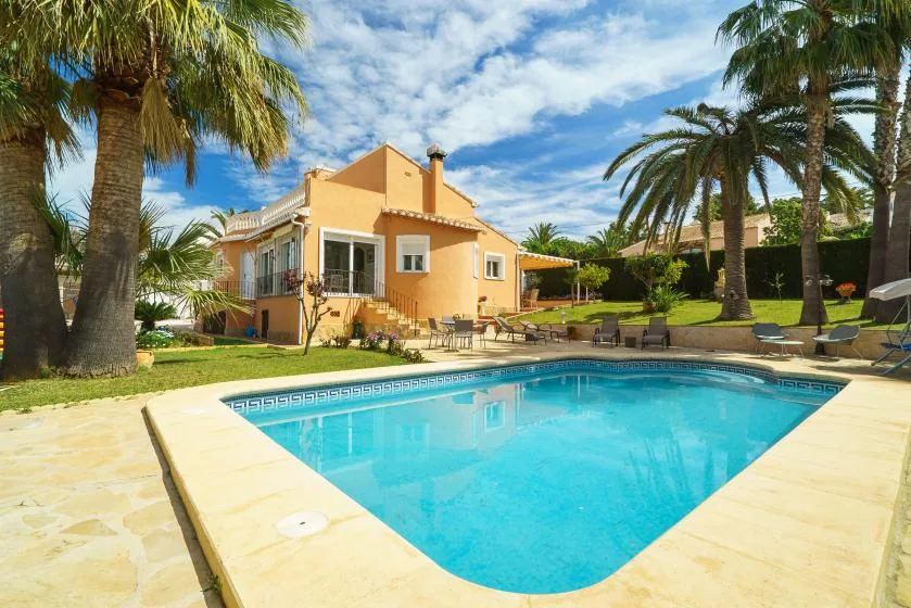 A swimming pool sits beside a yellow house and palm trees.