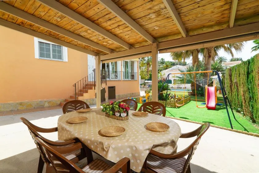 A covered patio has a round dining table, with a swing set visible in the yard.