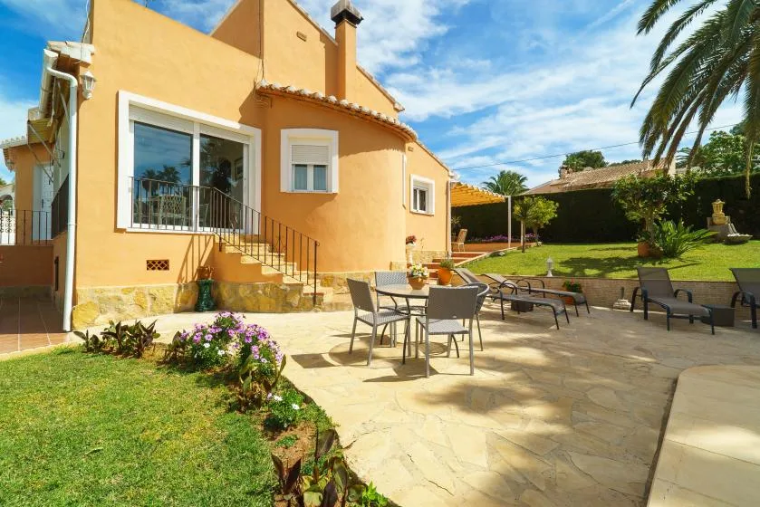 Un patio soleado junto a una casa amarilla, con asientos de jardín y césped.