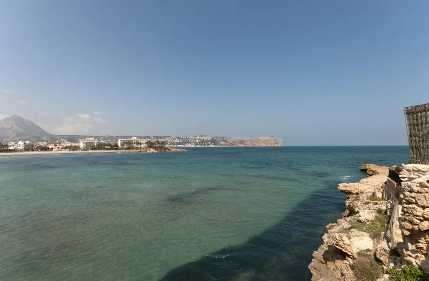 A coastal seascape with clear water, rocky shoreline, and distant buildings.