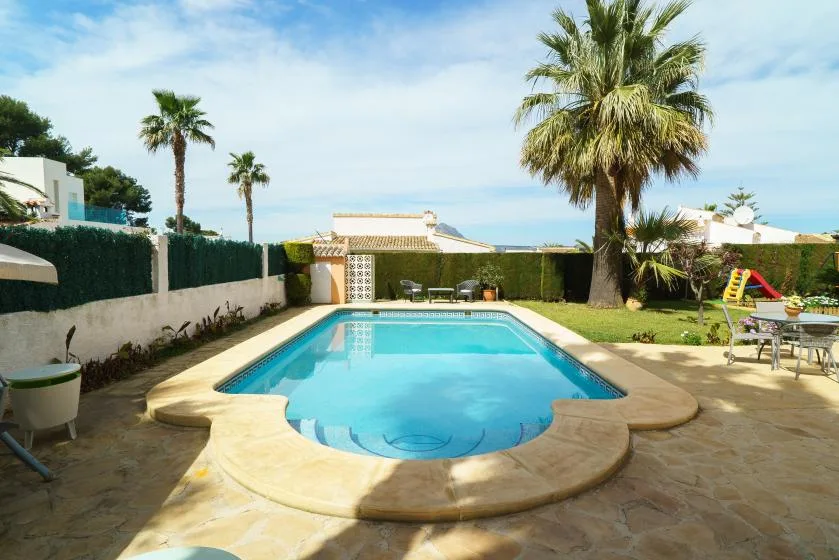 An outdoor swimming pool sits in a sunny yard with palm trees and patio seating.