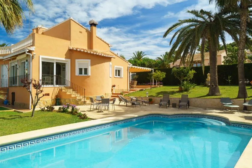 A swimming pool beside a yellow house, with patio seating and palm trees.