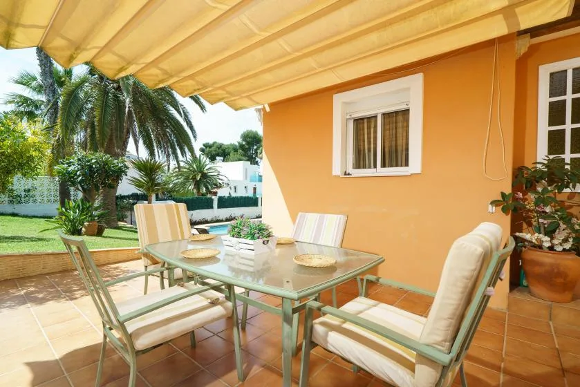 A shaded outdoor dining area with a glass table and cushioned seating.