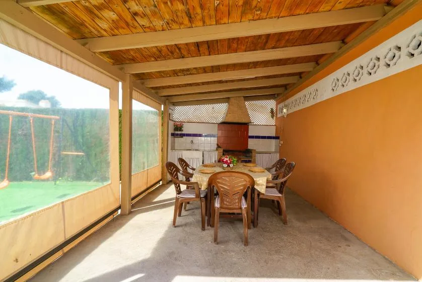 A covered patio has a long dining table surrounded by chairs.