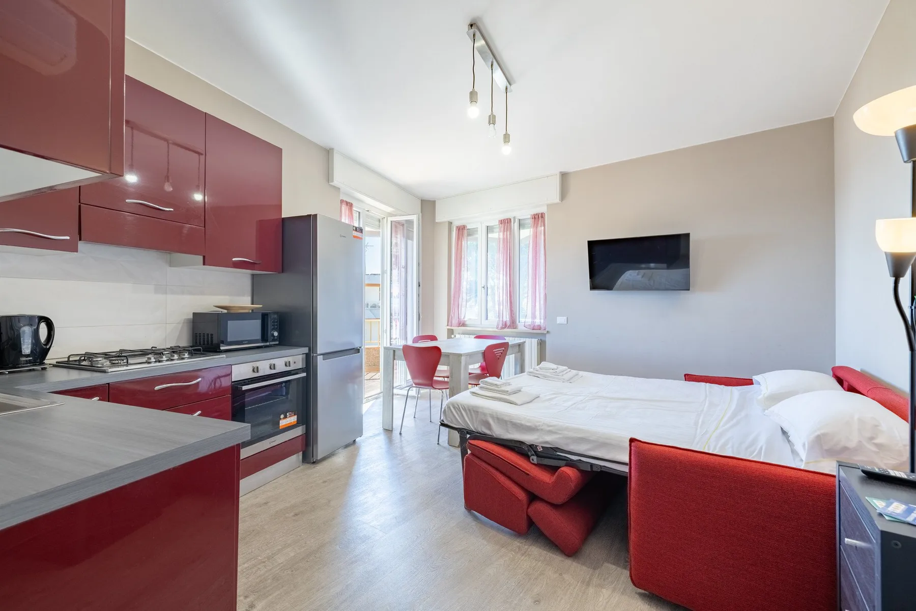 A bright studio living area combines a red kitchen, dining table, and bed.