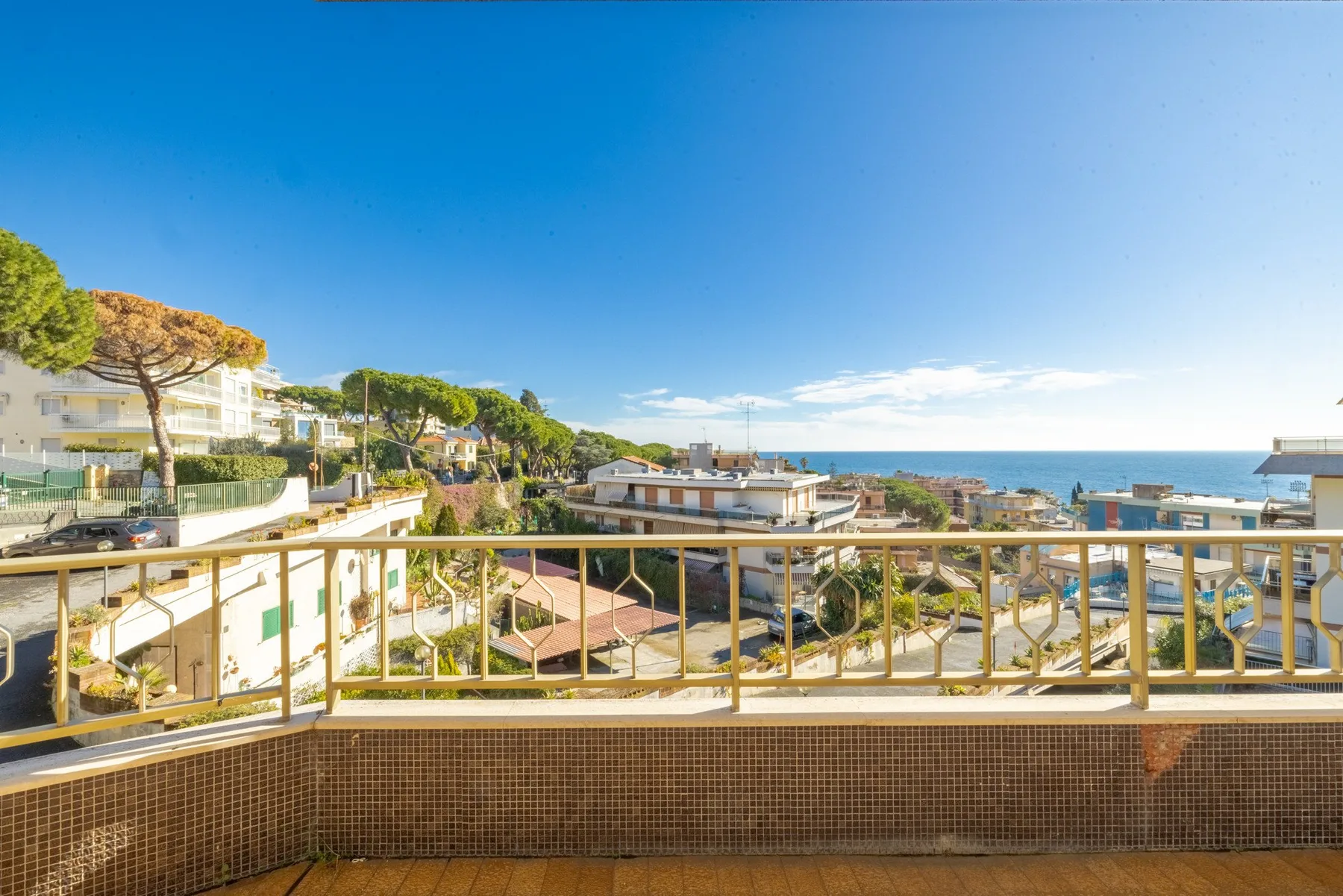 A balcony overlooks nearby buildings, trees, and the sea beneath a clear blue sky.