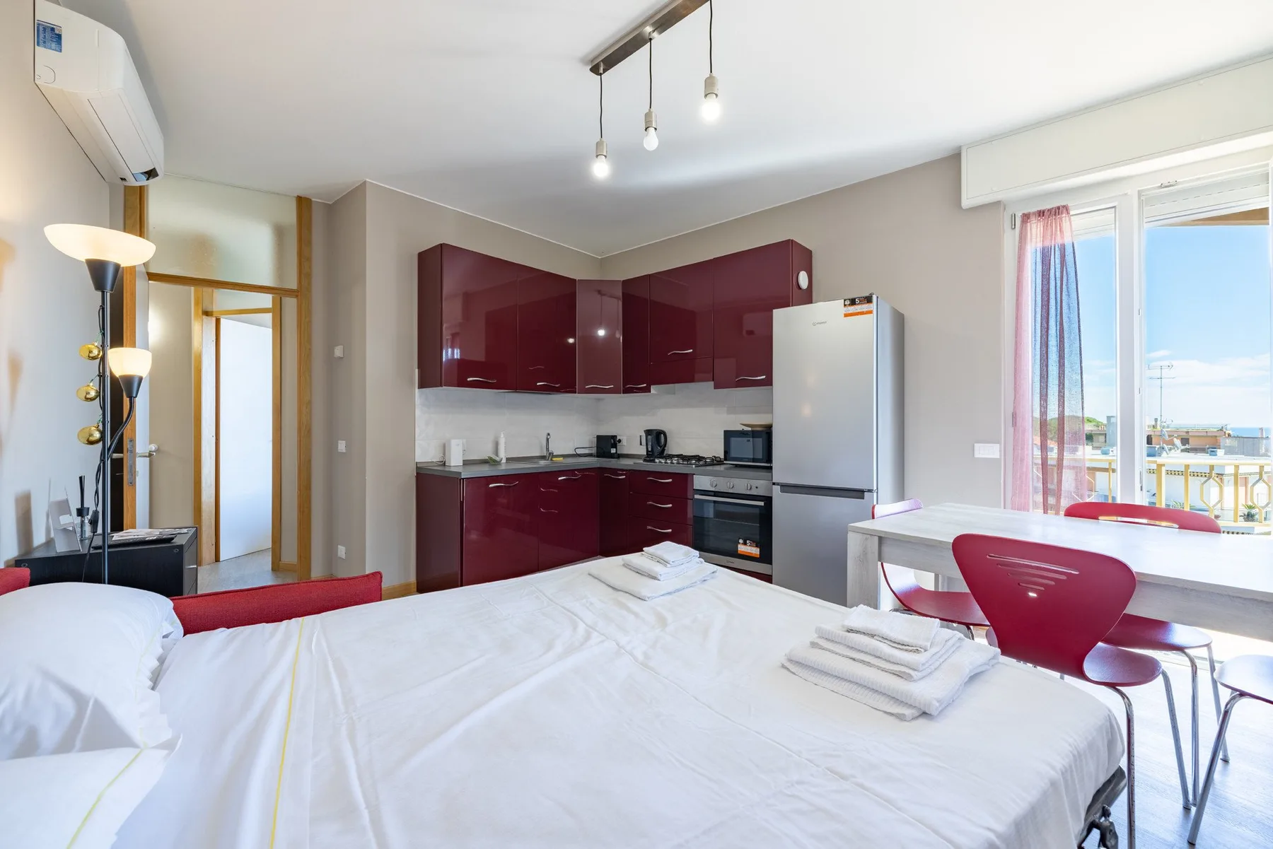 A bedroom with a bed, kitchenette, and dining table beside a bright window.