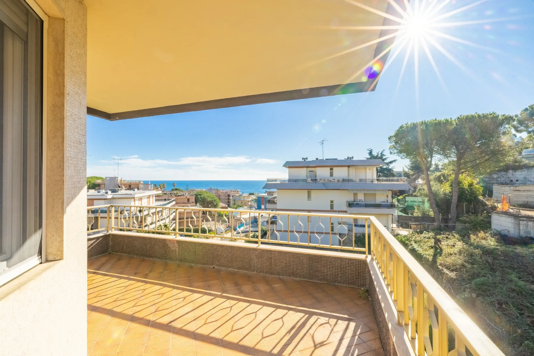 A sunny balcony overlooks nearby buildings, trees, and the sea.