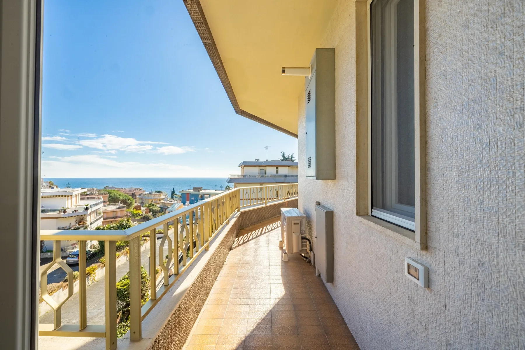 A sunlit balcony overlooks nearby buildings and the sea.