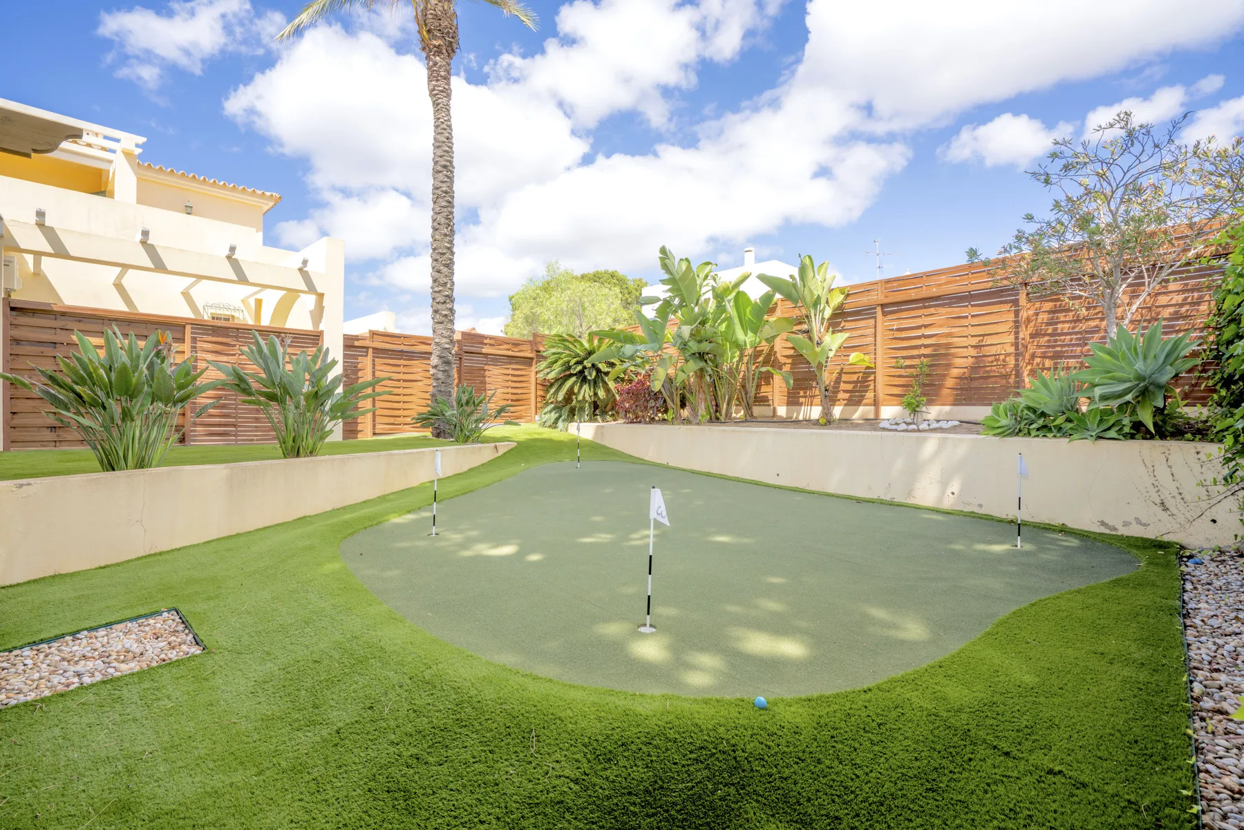 A small outdoor putting green sits among tropical plants and wooden fencing.