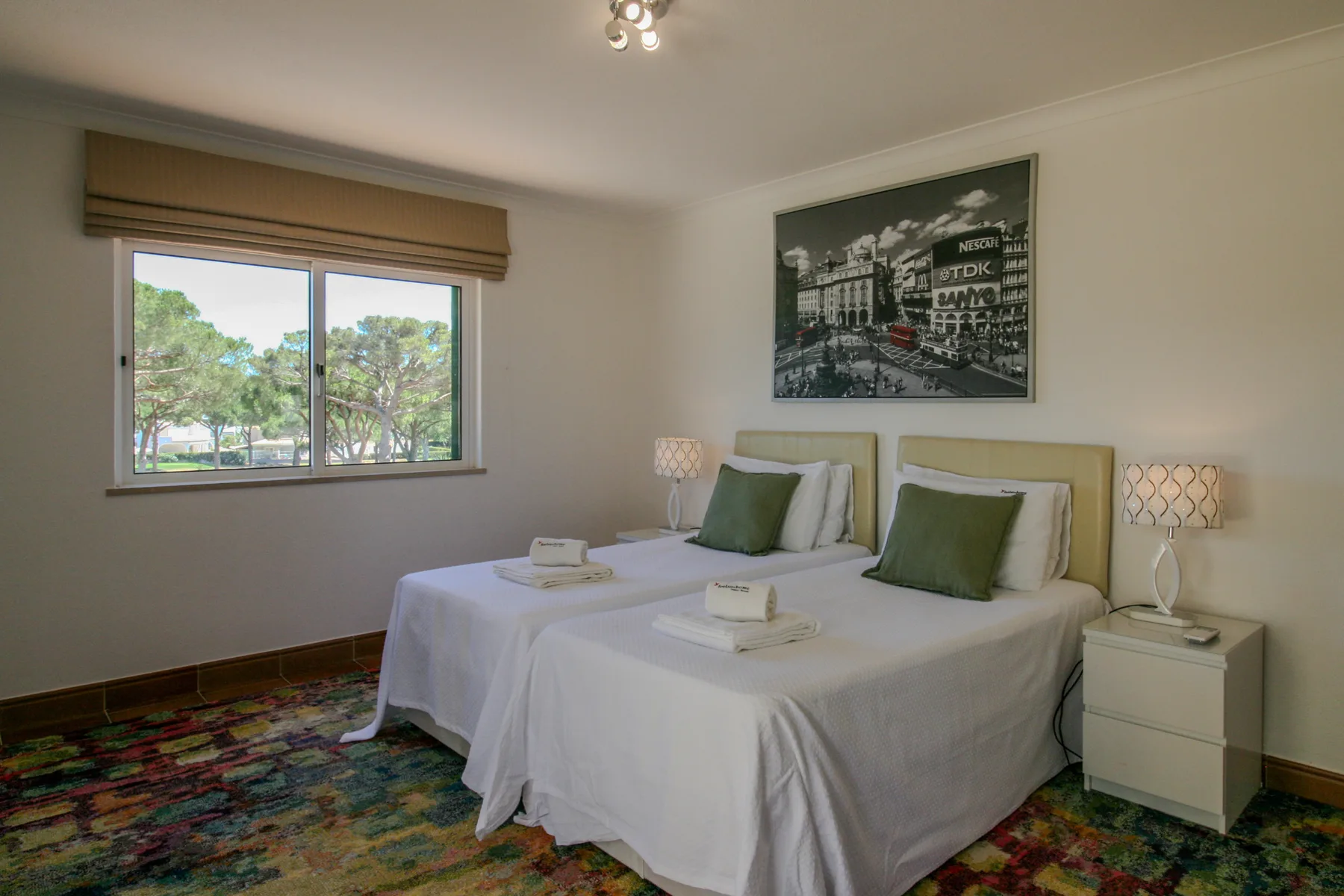A bedroom with two beds, bedside tables, and a window overlooking trees.