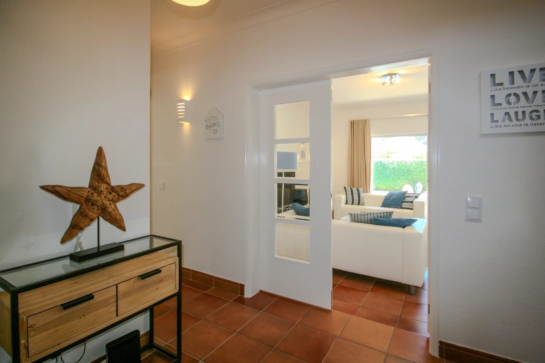 An interior hallway opens into a bright living room with seating and a window.