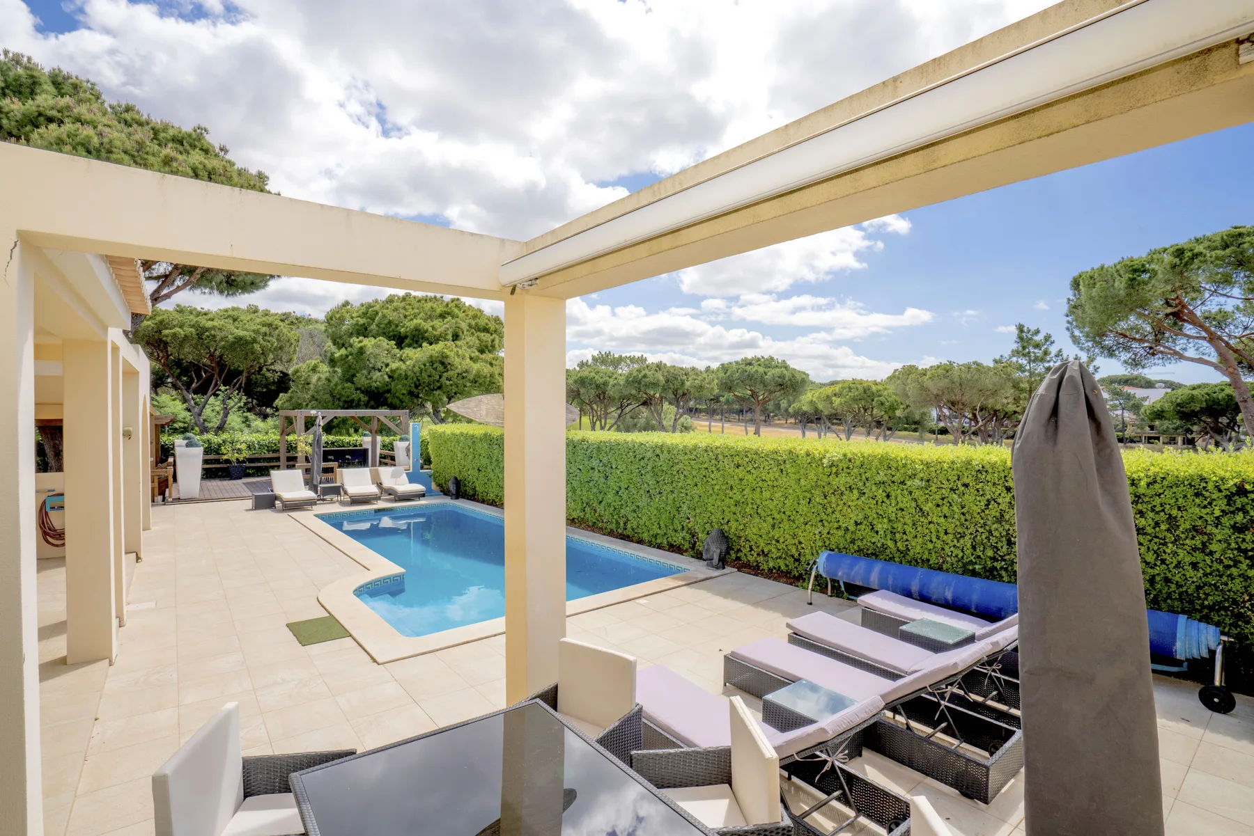 A tiled patio with a swimming pool, outdoor seating, and a hedge.