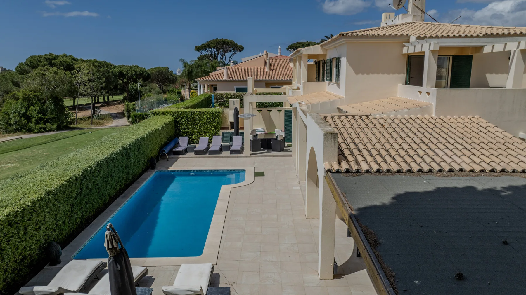 A villa patio overlooks a swimming pool bordered by lounge seating and hedges.