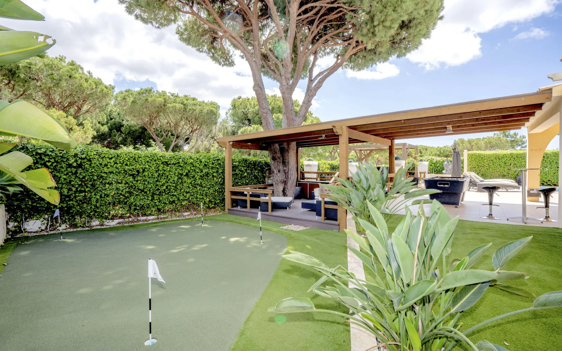 A garden patio with a wooden pergola, outdoor seating, and a small putting green.