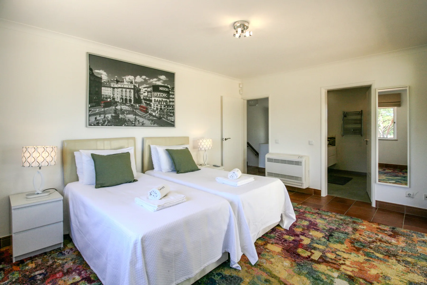A bright bedroom with two beds, bedside lamps, and a colorful patterned rug.