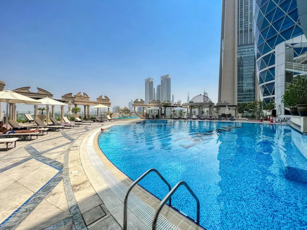 Una piscina curva junto a una terraza soleada con tumbonas y sombrillas.