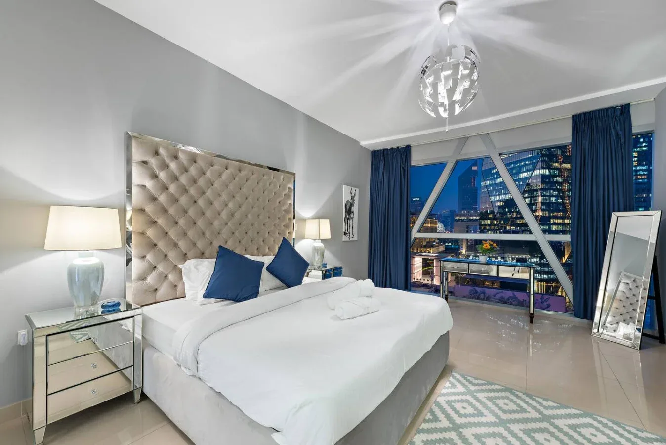 Un dormitorio con una cama blanca, cortinas azules y una gran ventana con vistas a las luces de la ciudad.