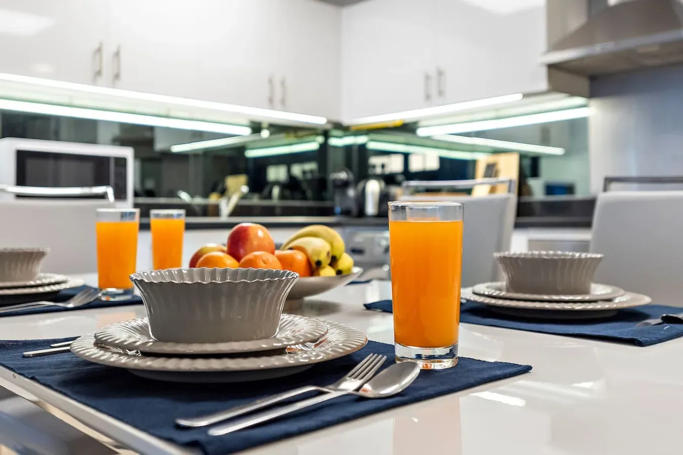 Una encimera de cocina está preparada con platos, cuencos, fruta y vasos de zumo de naranja.