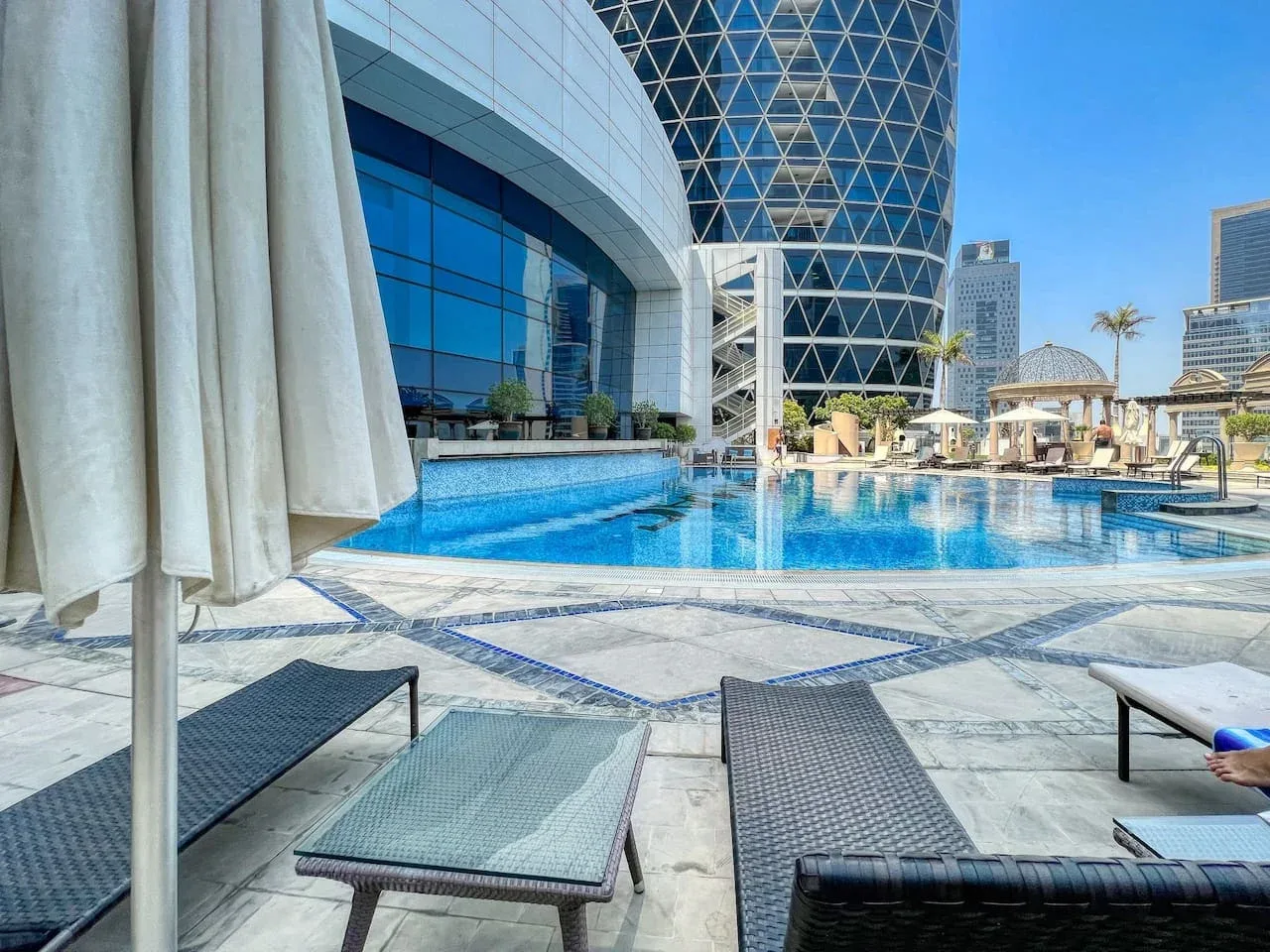 Una piscina exterior junto a un edificio moderno de vidrio, con asientos de descanso en la terraza.