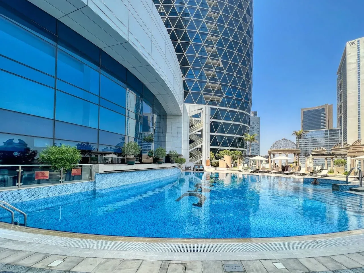 Una piscina en la azotea junto a edificios modernos de vidrio bajo un cielo despejado.