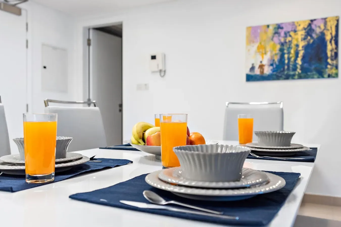 Una mesa de comedor está preparada con platos, bebidas, fruta y una pintura colorida en la pared.