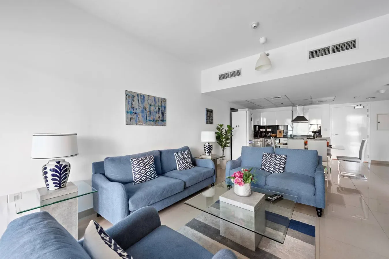 Una sala luminosa con asientos azules, mesas de vidrio y una cocina abierta al fondo.