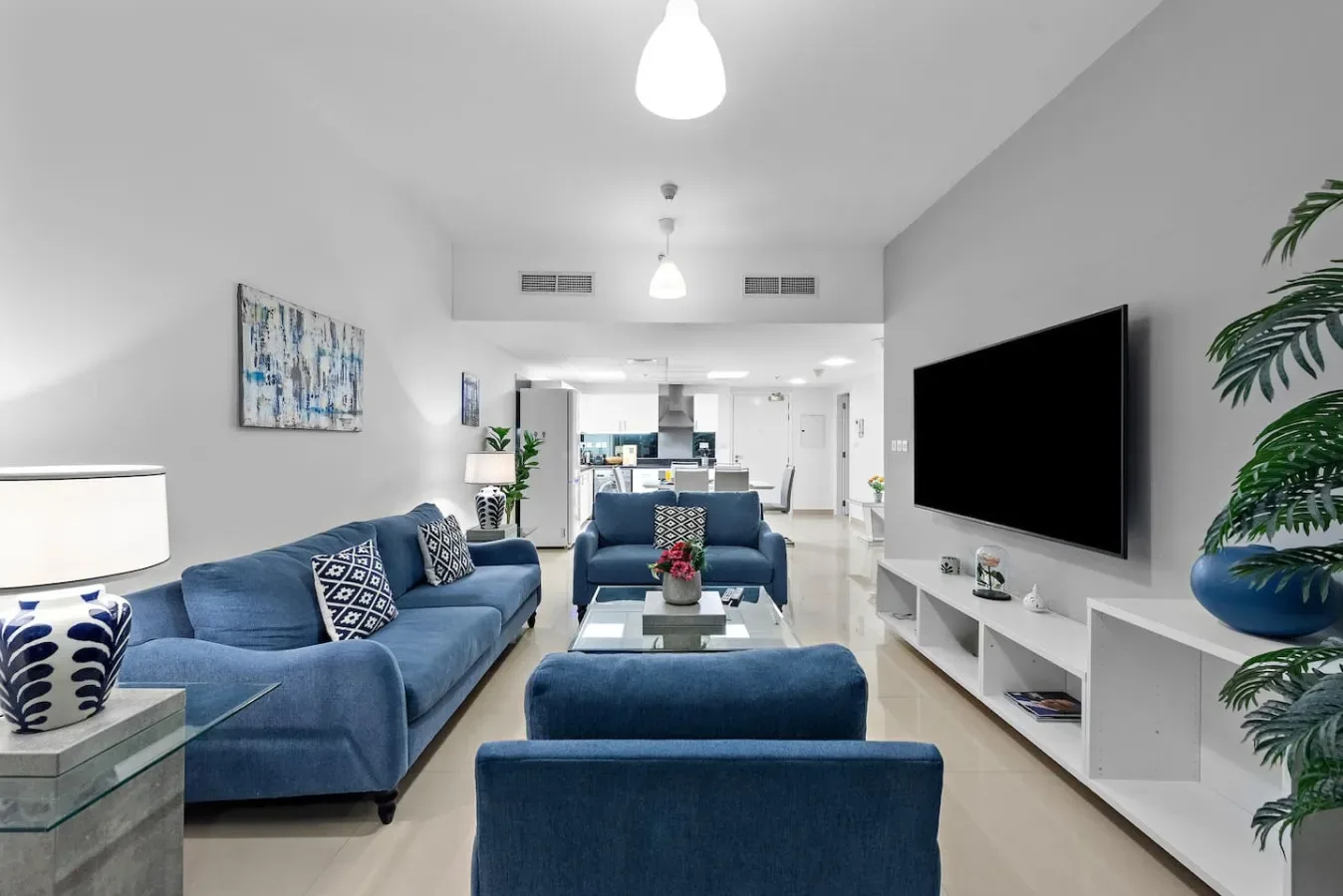 Una sala de estar luminosa con asientos azules, un televisor en la pared y una cocina abierta al fondo.